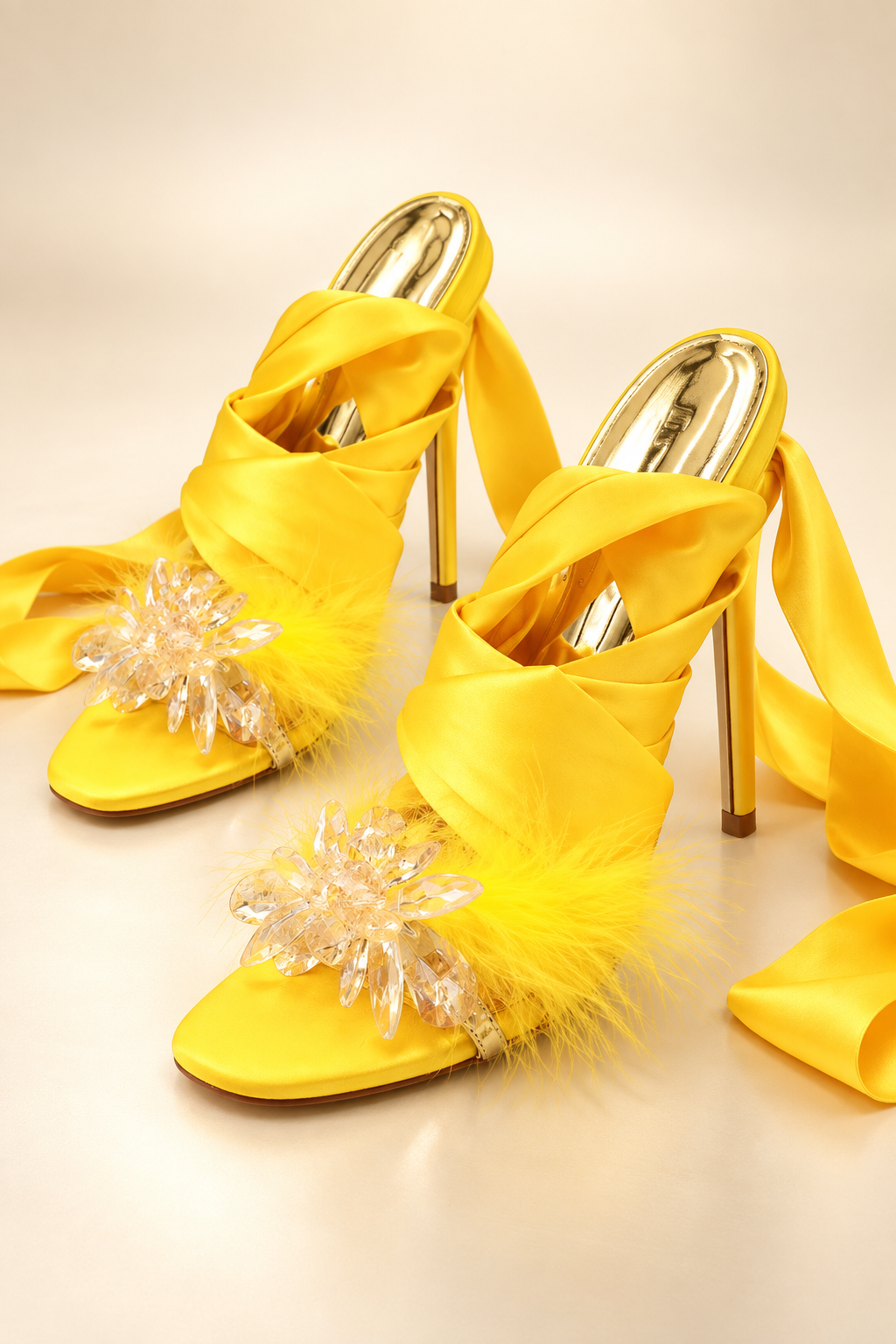 Golden Hour vivid yellow satin cross-strap stiletto heels on a warm neutral studio background, showing crystal-flower toe ornaments, yellow feather trim, gold footbeds, long wrap ribbons and slim gold heels.
