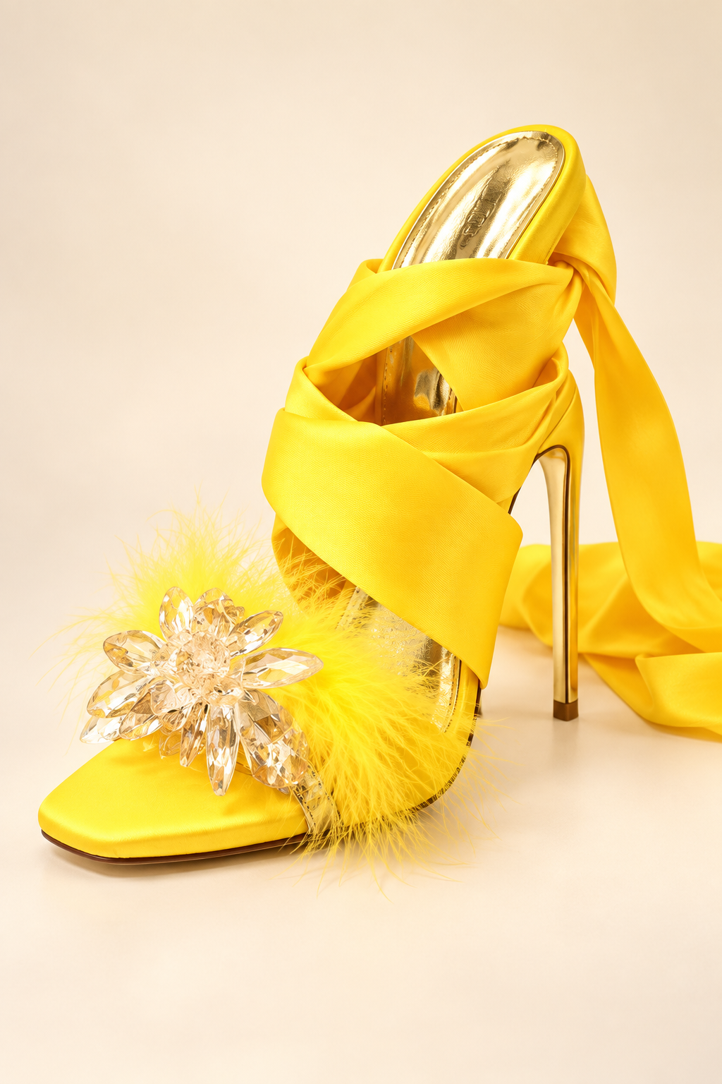 Golden Hour yellow satin stiletto heel close-up showing the clear crystal-flower toe ornament, feather trim, square open toe, cross straps, long wrap ribbon, reflective gold footbed and slim gold heel.