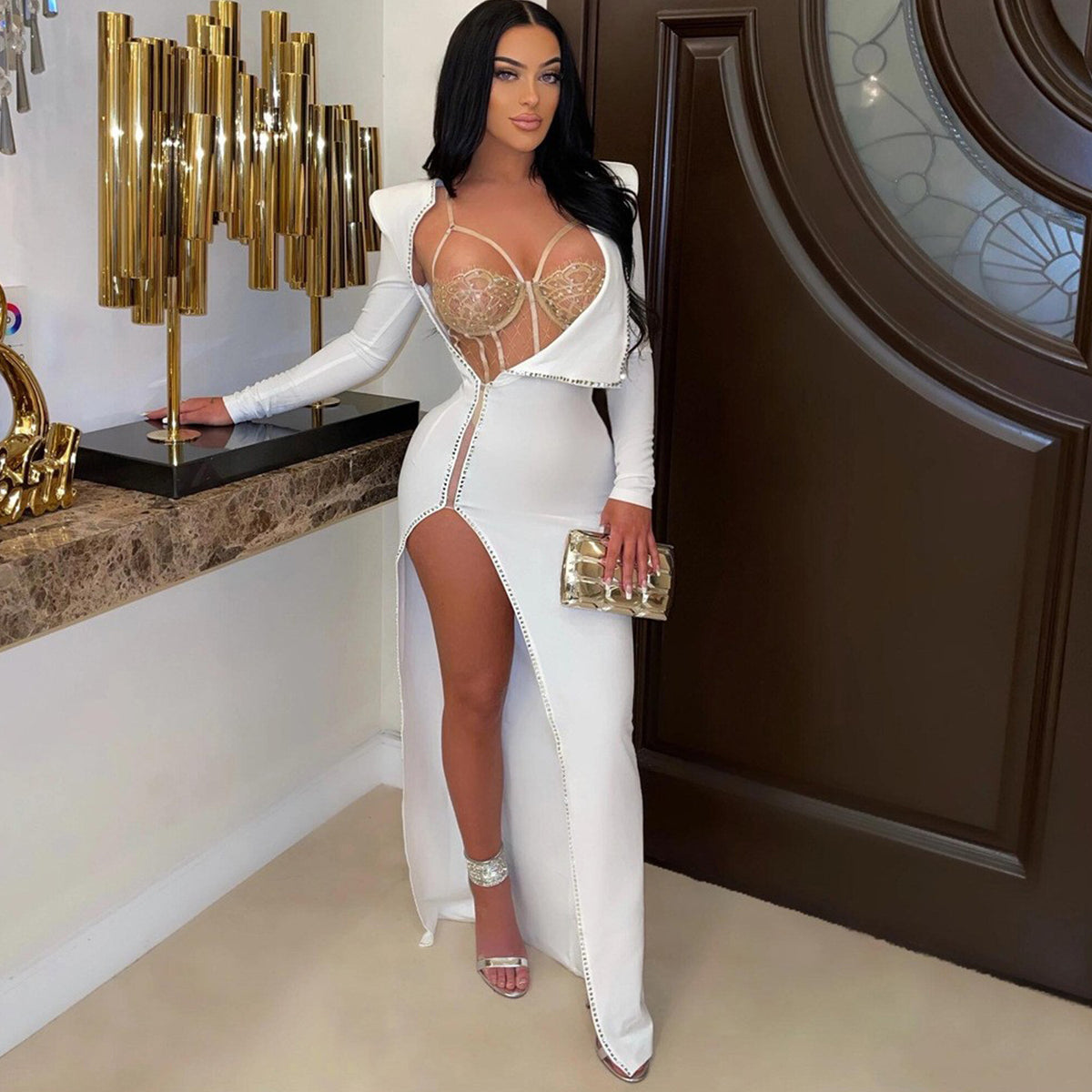 Model wearing the white Billion Moves Maxi Dress with long sleeves, jeweled sheer bodice, high thigh slit, and matching clutch in an elegant interior.