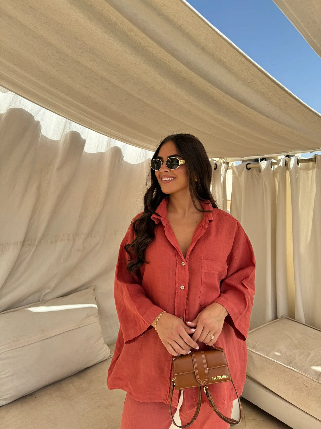 Front three-quarter view of an adult model wearing the orange-red shirt and trouser set with sunglasses and a handbag.