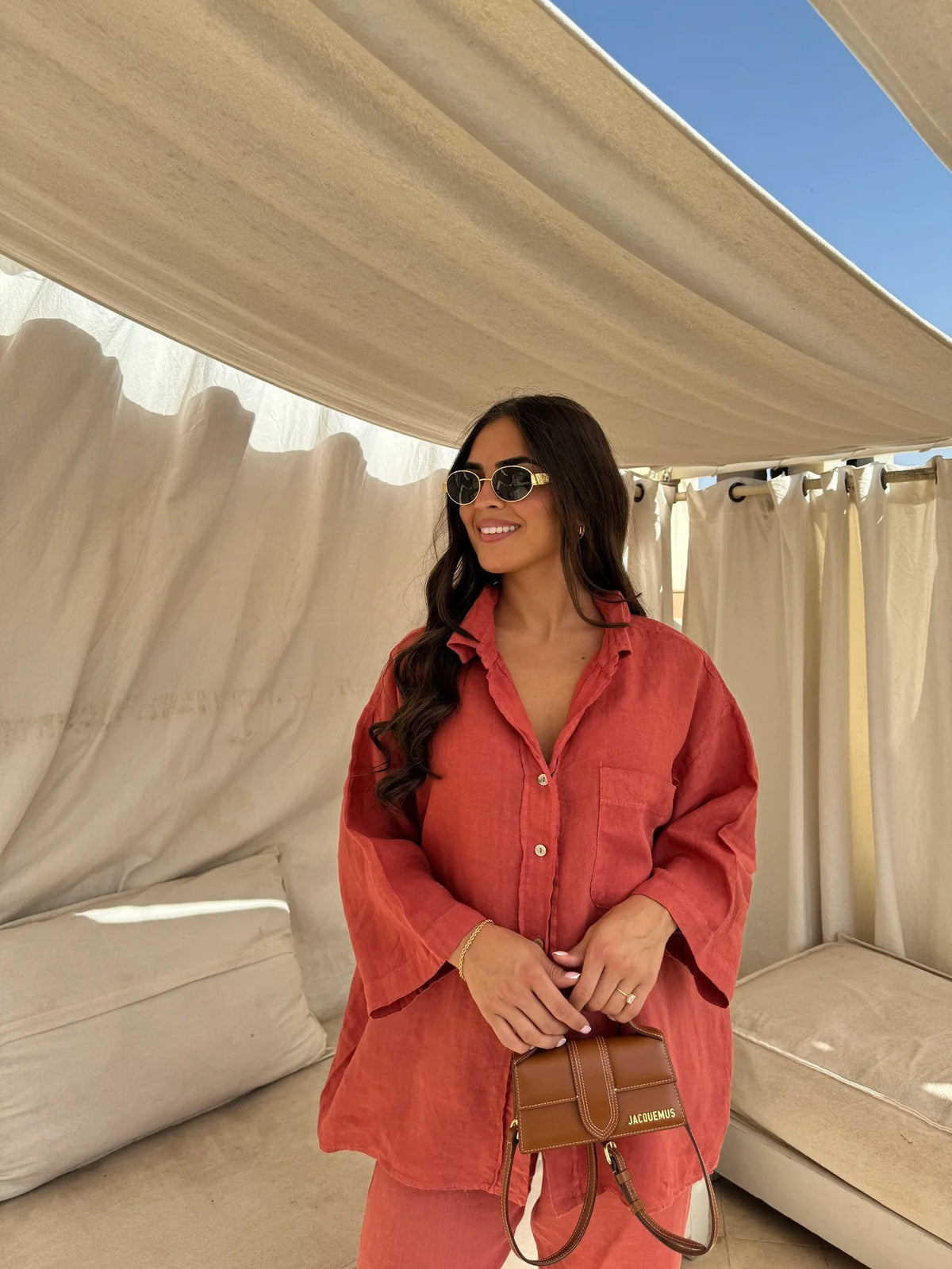 Front three-quarter view of an adult model wearing the orange-red shirt and trouser set with sunglasses and a handbag.
