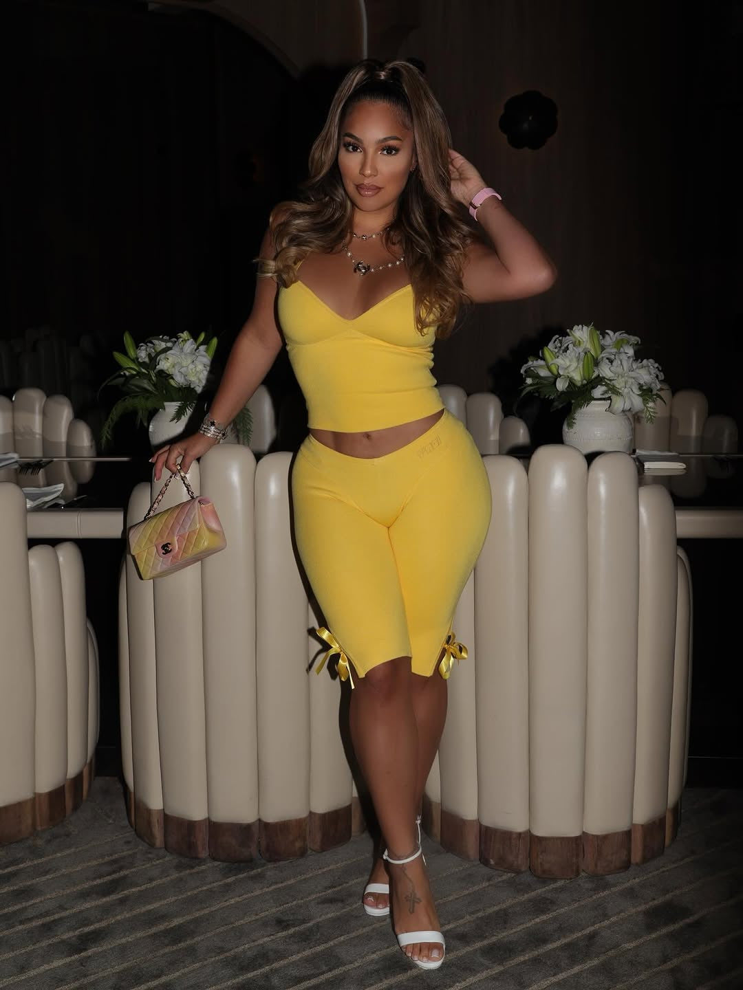 Full-length view of a model wearing a yellow cropped top and yellow side-tie cropped pants while holding a handbag in a lounge.