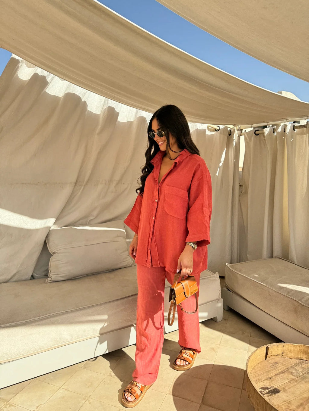 Full-length view of an adult model wearing the orange-red shirt and trouser set on a shaded patio.