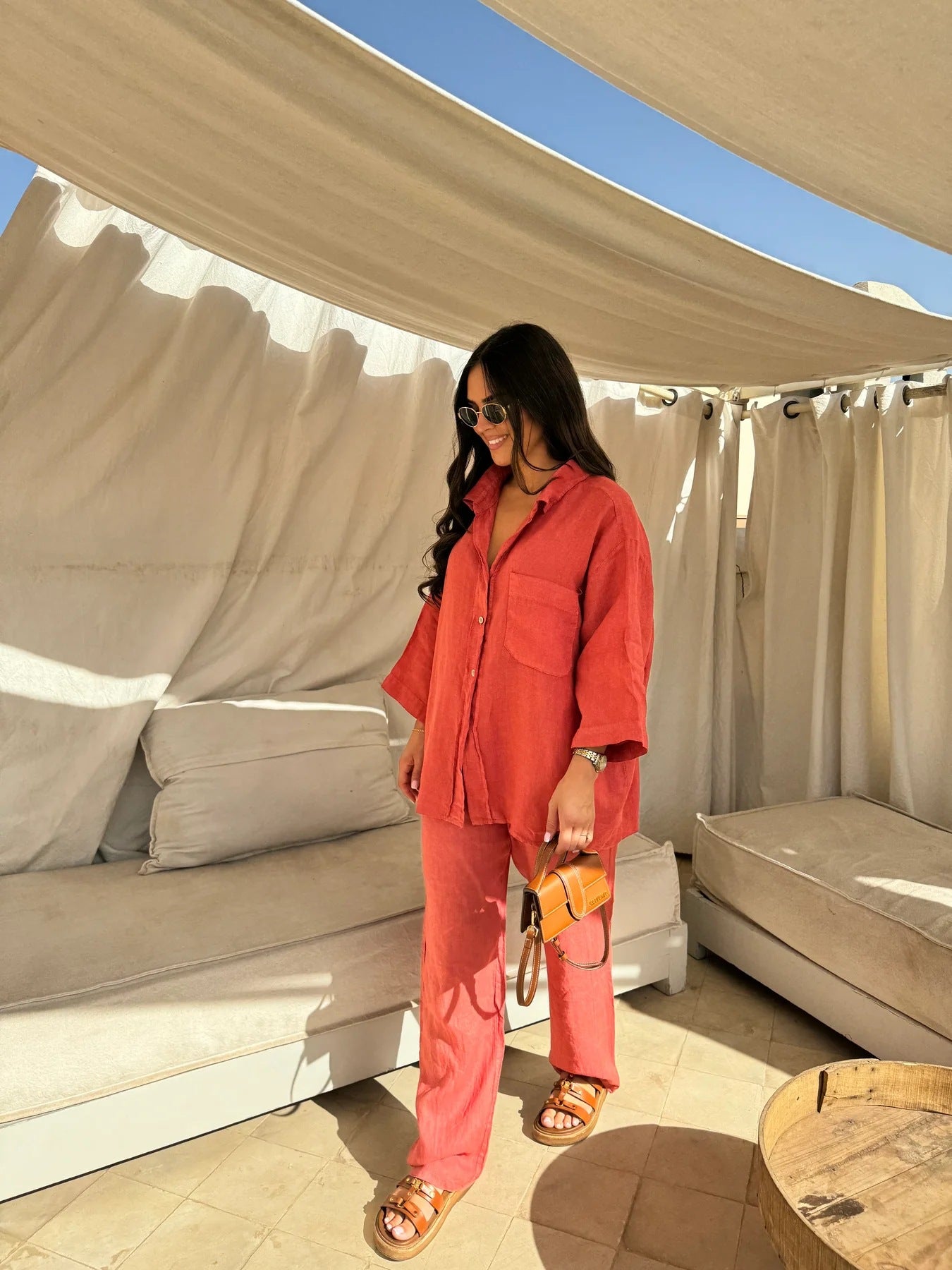 Full-length view of an adult model wearing the orange-red shirt and trouser set on a shaded patio.